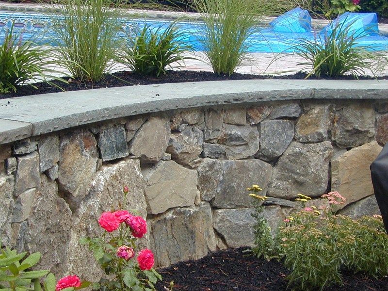 A curved stone retaining wall topped with gray capstones, fronted by pink roses and backed by decorative grasses and a pool.