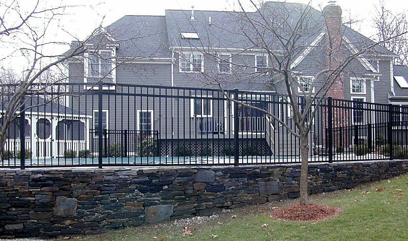 A multi-story gray house with a black metal fence surrounding a pool above a stone retaining wall.