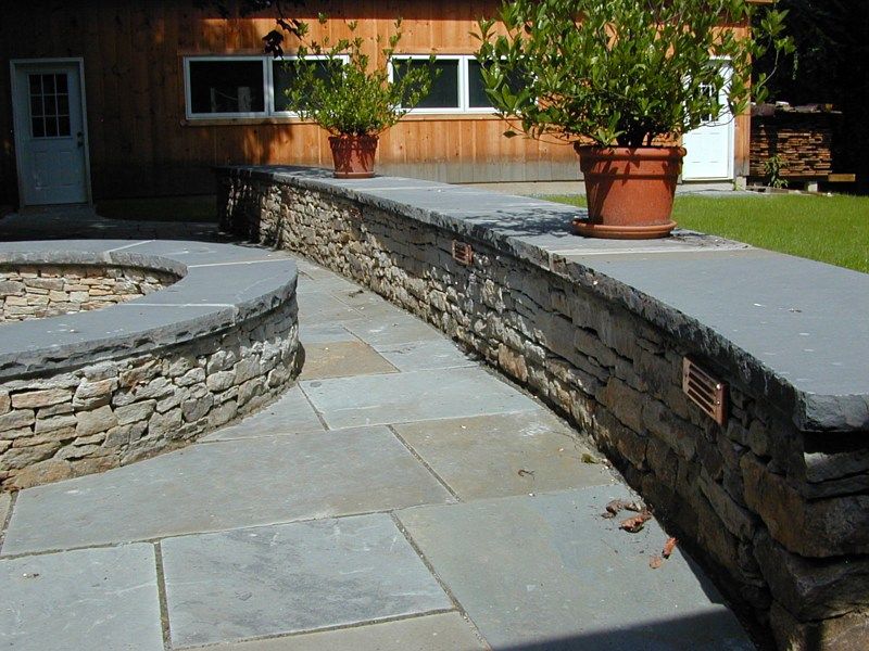 A curved stone retaining wall and patio with bluestone caps, potted plants, and built-in lighting in front of a house.