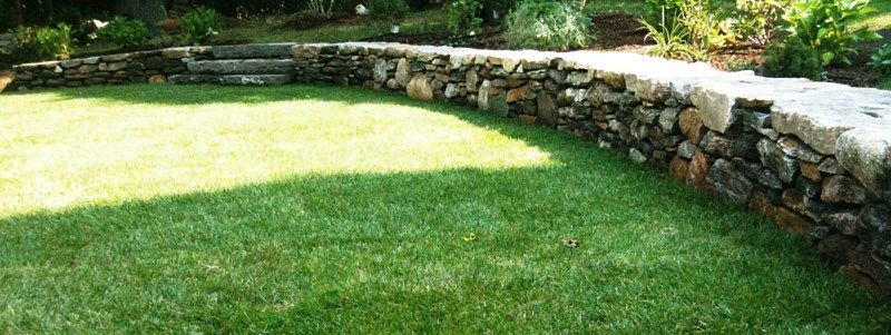 A low, natural stone retaining wall curves through a lush green lawn, separating the grass from a slightly raised garden.