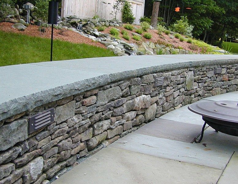 A curved stone retaining wall with a smooth gray cap and integrated lighting, bordering a patio and a landscaped hill.