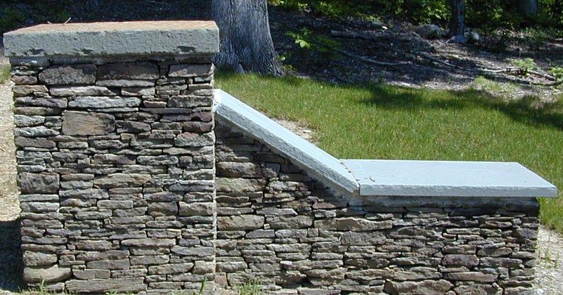 A stone pillar with an attached low retaining wall topped with light gray concrete caps, set against a grassy background.