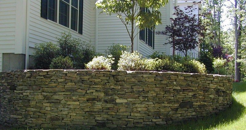 A curved, stacked-stone retaining wall sits in front of a white house with landscaping.