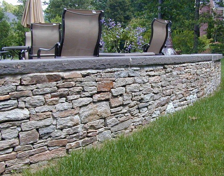 A low, curved stone retaining wall topped with a smooth concrete cap borders a patio with outdoor seating on grass.