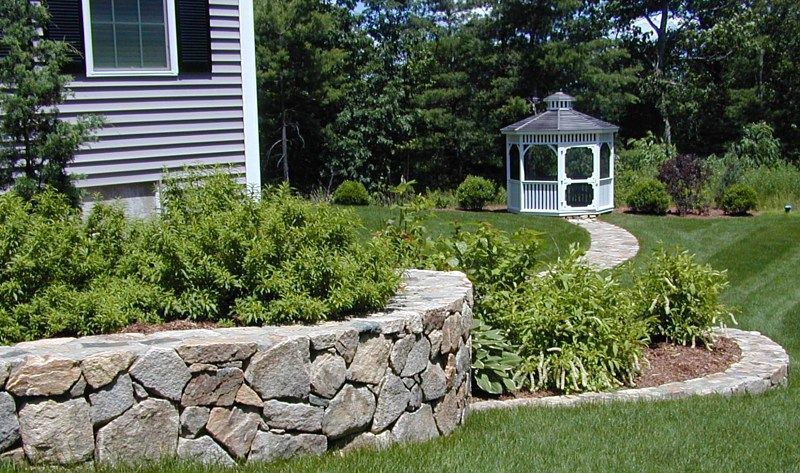 A curved stone retaining wall borders a garden bed, with a stone path leading to a white gazebo in a grassy backyard.