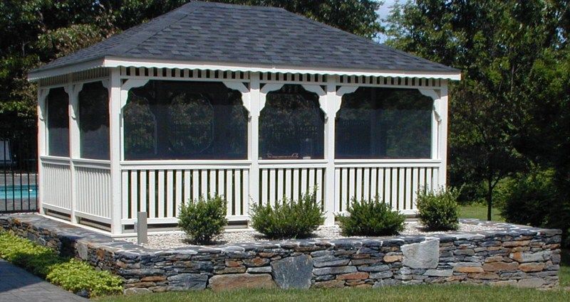A white screened-in gazebo with a dark shingled roof, set on a stone wall terrace next to a pool.