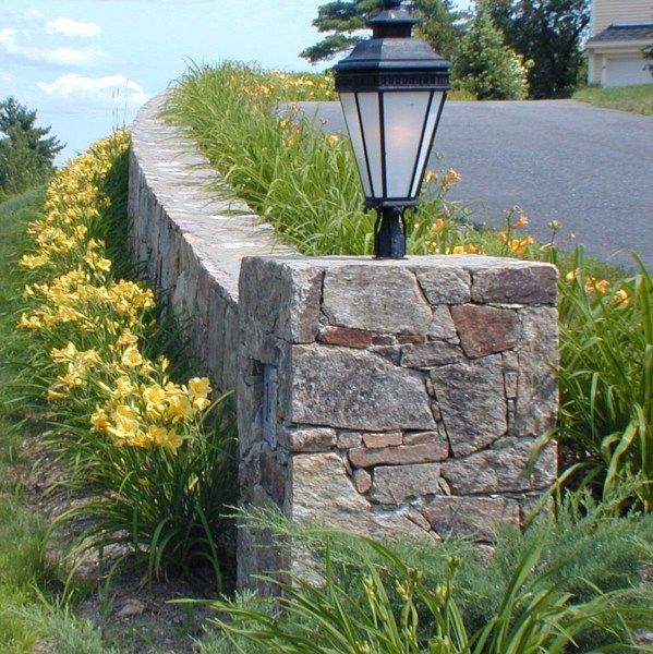 A stone wall topped with a black lantern lamp, bordered by yellow daylilies along a curved driveway.