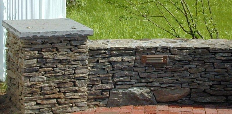 A stone pillar and wall featuring a built-in recessed outdoor light, set against a background of grass and a white fence.