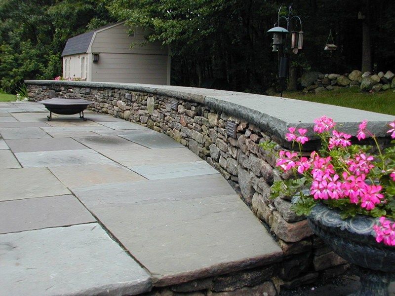 A stone patio with a curved retaining wall, a fire pit, and a planter with bright pink flowers in a garden setting.