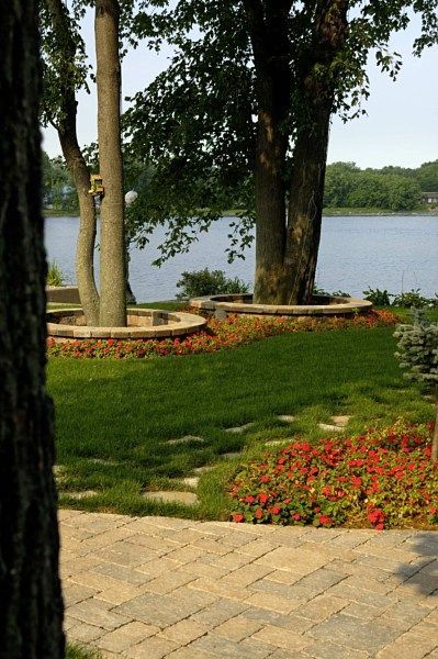 A lakeside garden features two trees with stone-walled planters, red flowers, a green lawn, and a stone patio.