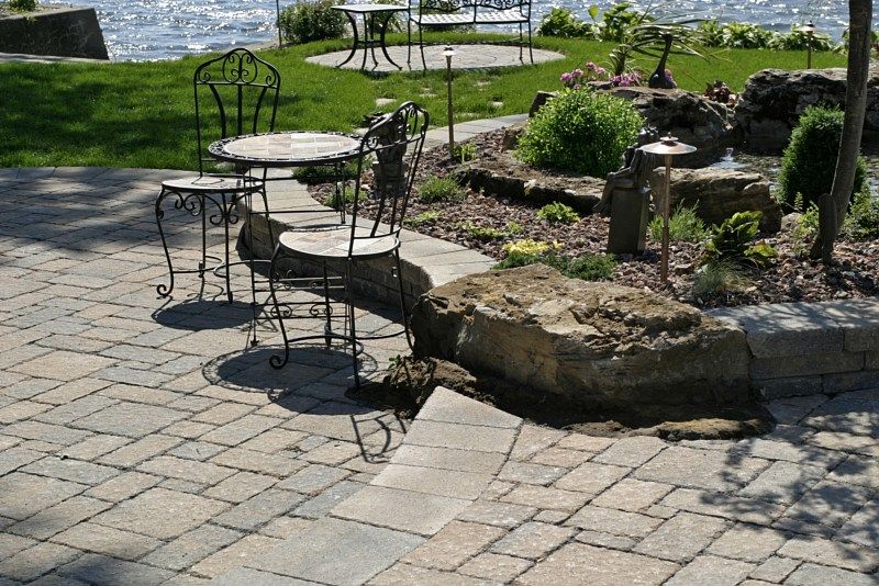 Stone patio with a wrought iron bistro set, large rocks, and landscaping overlooking a lake.