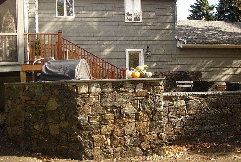 A stone outdoor kitchen island with a covered grill sits on a patio next to a gray house with a wooden deck.