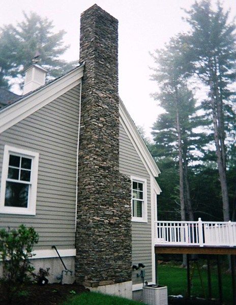 A gray house with white trim features a tall, exterior stone chimney, next to a raised wooden deck and tall pine trees.