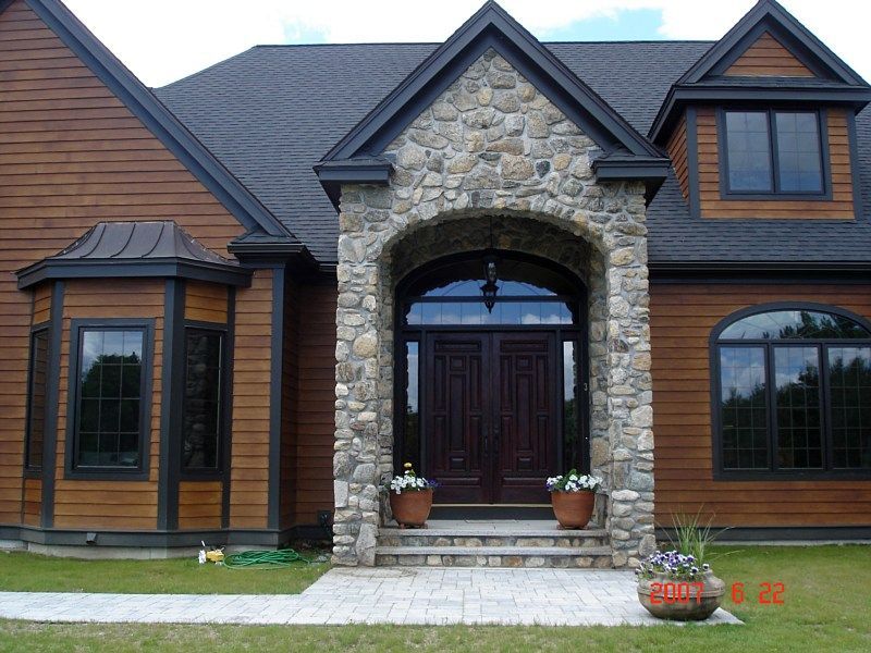 A house with horizontal wooden siding, a dark roof, and a prominent stone entryway featuring a double door and arch.