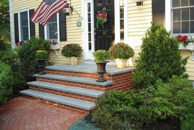 A front entrance with brick steps and a stone walkway leading to a black door on a yellow house, flanked by shrubs.