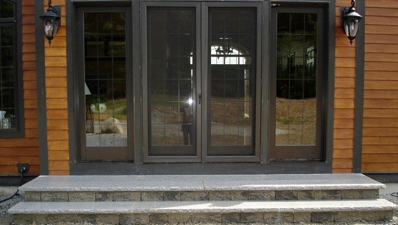 Two wide stone steps leading up to a pair of sliding screen doors on a wood-sided house with two exterior lamps.