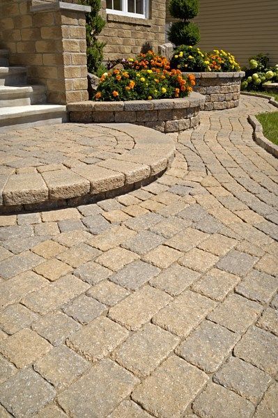 A curved stone paver walkway and circular step lead to a house entrance with raised flower beds and a stone pillar.