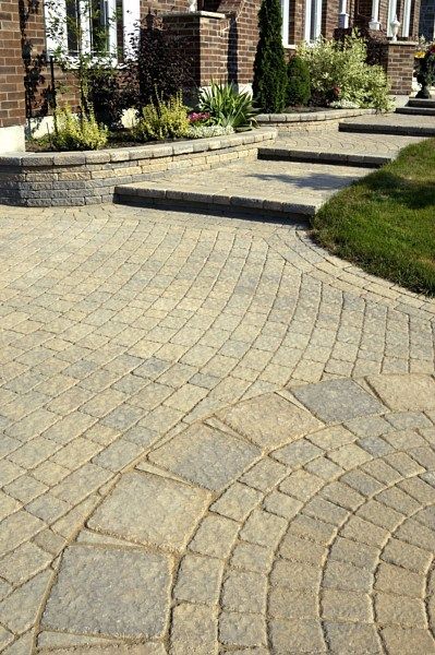 A tan and grey stone paver patio and walkway leading to stone stairs and a brick house entrance.