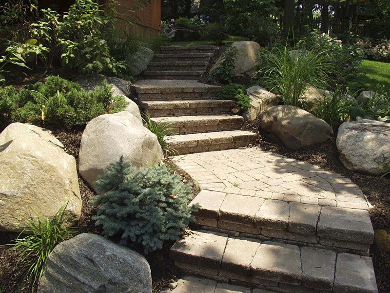 Stone steps with a paver walkway leading up a landscaped garden path surrounded by large boulders and green shrubs.
