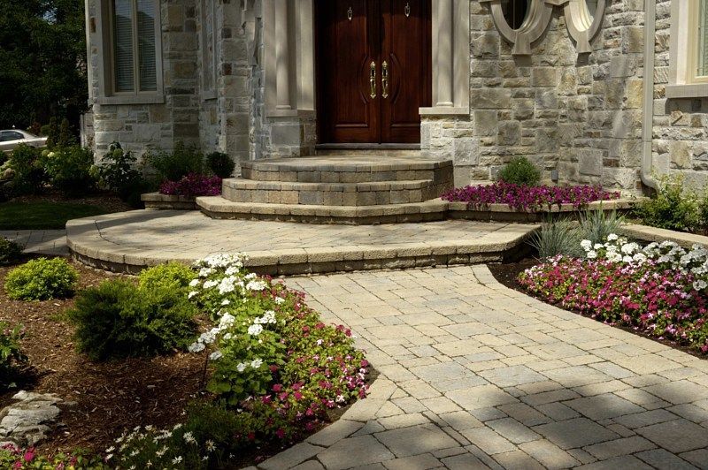 A stone walkway leads to a curved front porch entrance of a house, framed by manicured gardens and vibrant flower beds.