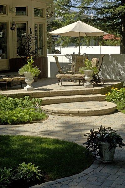 A sunny patio with paved steps, outdoor furniture, a white umbrella, and potted plants next to a house with large windows.