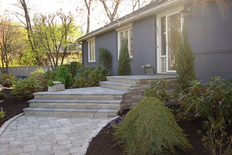 Stone steps and a paved walkway leading to the entrance of a dark-gray brick house, surrounded by landscaped shrubs.