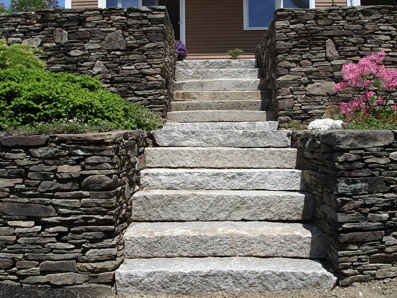 Granite steps lead up to a house entrance between two dry-stacked stone retaining walls with greenery and pink flowers.