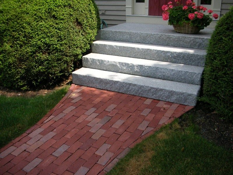 A brick path leads up to a set of four light gray granite steps in front of a house entrance with a potted flower.