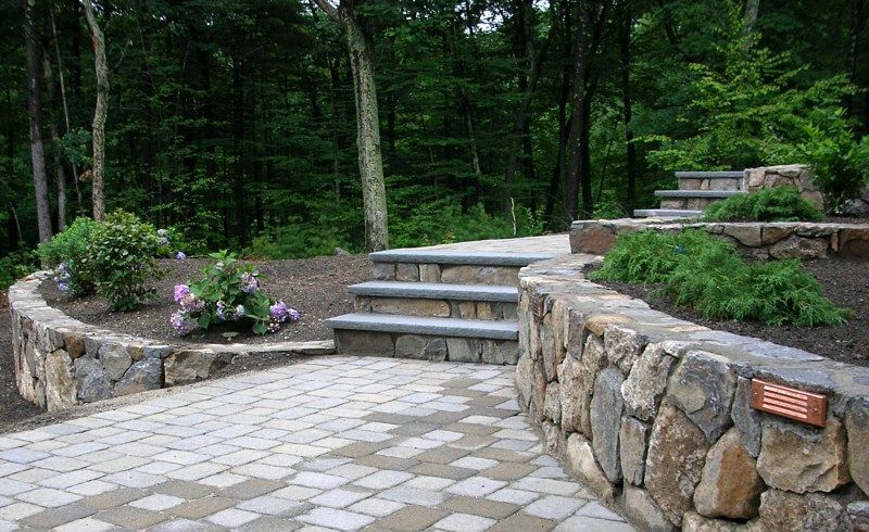 A stone patio features a curving retaining wall, a paved walkway, and steps leading up into a wooded landscape.