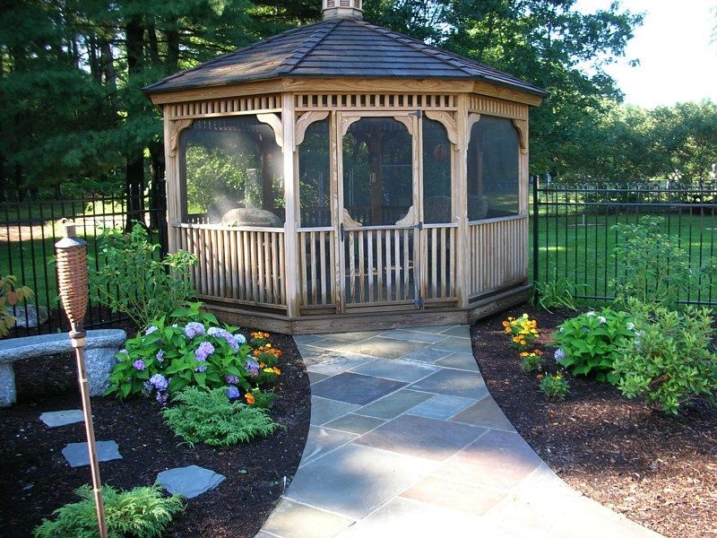 A wooden gazebo with screened panels sits at the end of a stone walkway in a garden with landscaping and a metal fence.