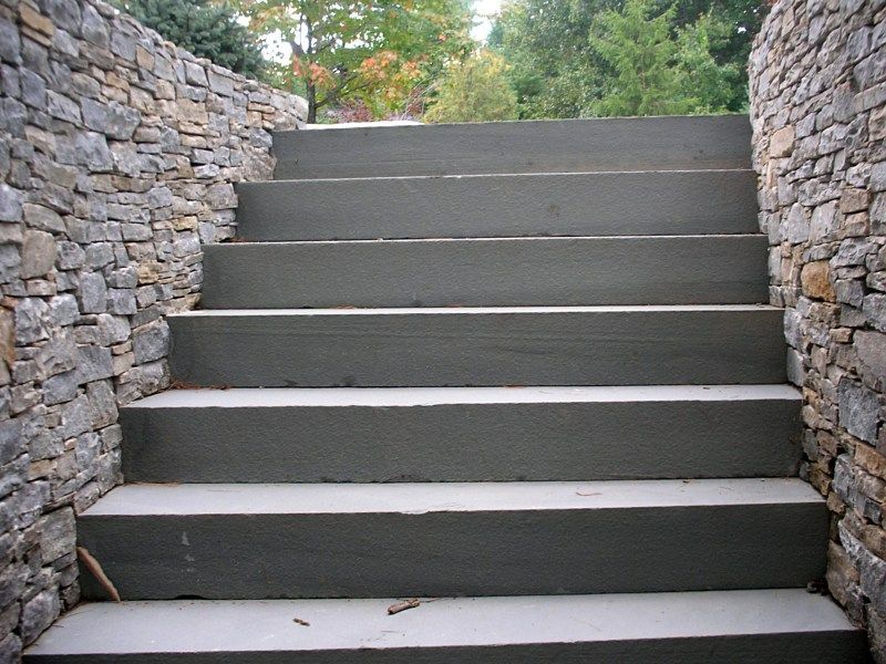 A straight set of wide gray concrete steps flanked by two textured stone walls with trees in the background.