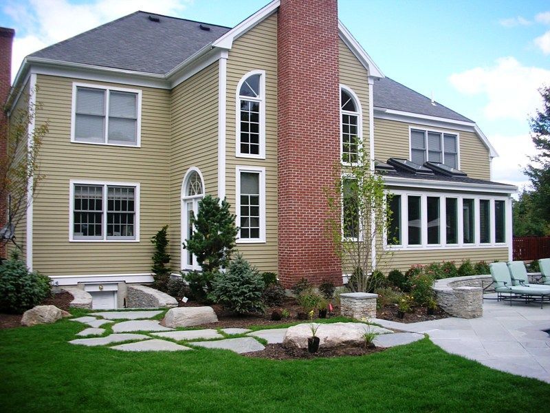 A tan-sided house with a large brick chimney, stone patio, and landscaped yard.