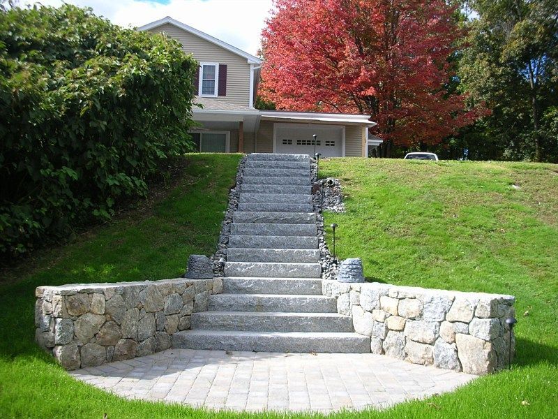 Stone steps lead up a grassy hill to a house with a brick patio at the base framed by low, stacked-stone walls.