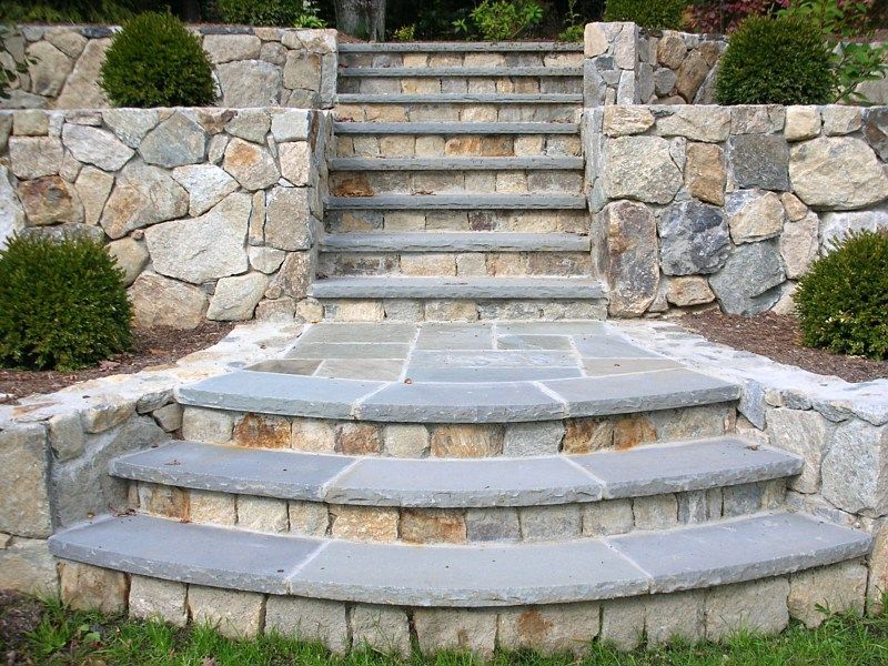 Outdoor stone steps with gray treads leading up a terraced garden wall with two rounded green shrubs.
