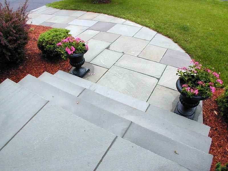Gray stone steps leading down to a flagstone walkway with two decorative planters of pink flowers in a garden setting.