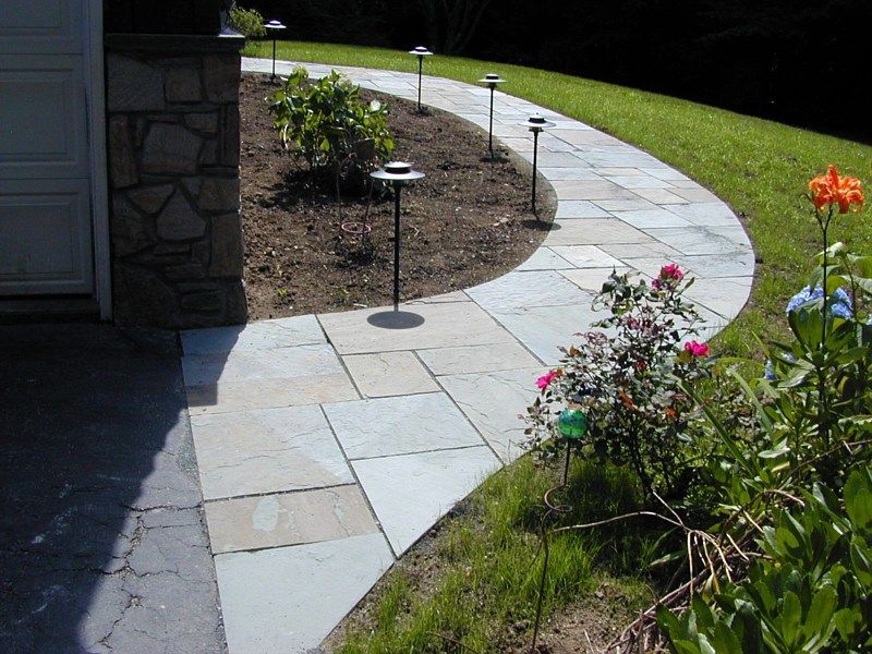 A curved stone walkway leads past a garden bed with landscape lighting next to a stone-faced garage.