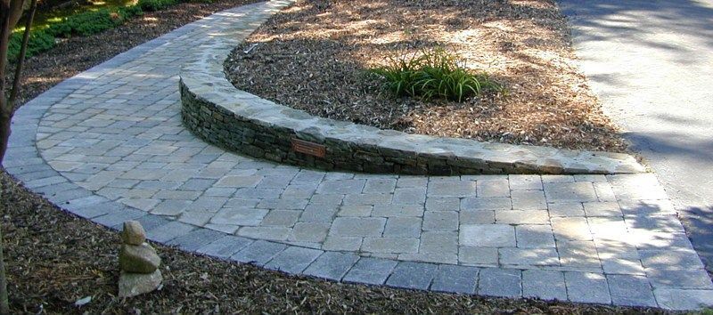 A curved stone walkway borders a mulch garden bed with a low stone retaining wall in a landscaped outdoor setting.