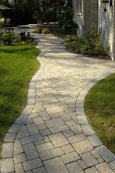 A winding stone paver walkway framed by grass leads toward a light-colored brick building in a landscaped garden.