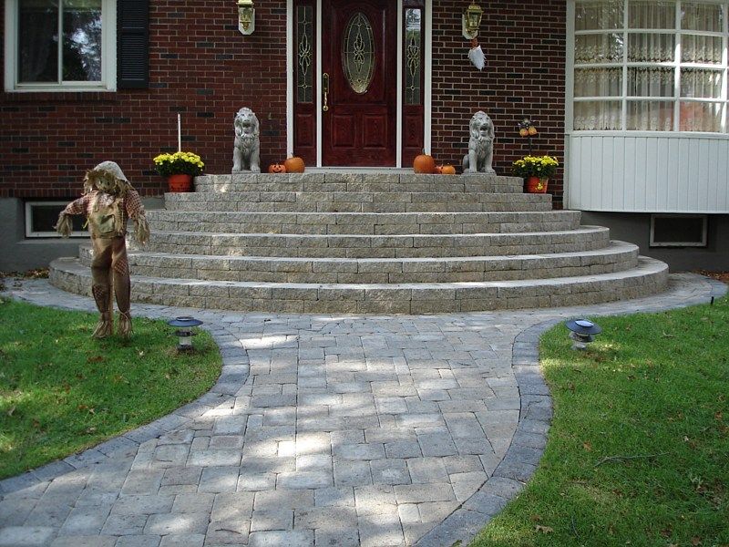 A curved stone paver walkway leads to a multi-tiered stone staircase at a brick home entry decorated for autumn.