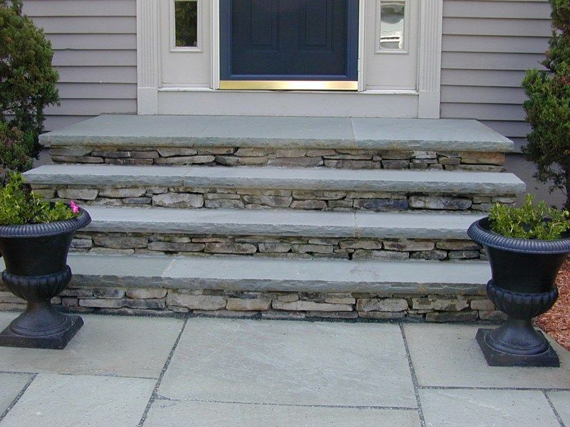 Four stone steps with flat blue-gray treads lead up to a navy blue front door, flanked by black planters on a patio.
