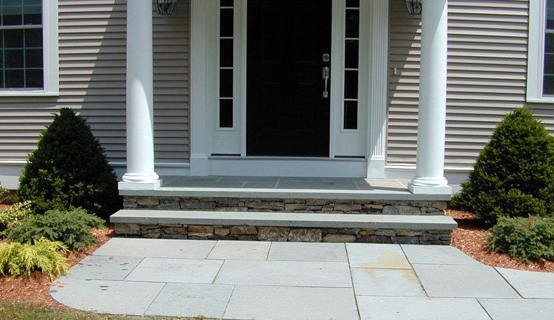 Front entrance of a house with white columns, stone steps, and a blue stone patio flanked by two evergreen bushes.