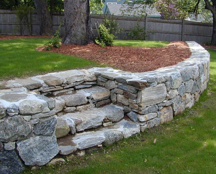 A dry-stacked stone retaining wall with built-in natural stone steps, set in a grassy yard near a tree and mulch bed.