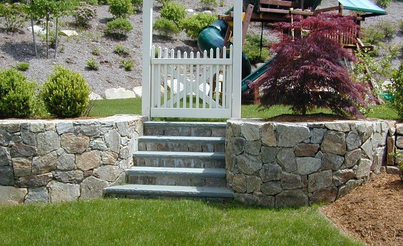 Stone retaining wall with a central set of stairs leading to a white picket gate in a backyard garden setting.