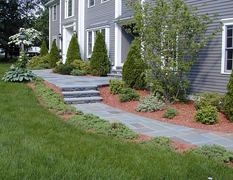 A stone walkway with two steps leads to a house with gray siding and landscaping featuring shrubs and red mulch.