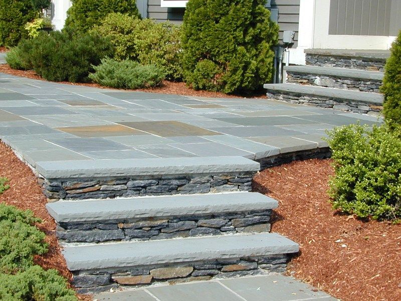 A stone patio with a set of three stone steps leading down to a lower level, surrounded by landscaping and wood mulch.