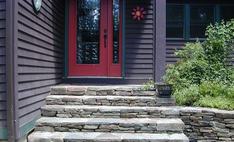 Stone steps lead to a red front door of a dark-sided house with a small planter and bushes nearby.