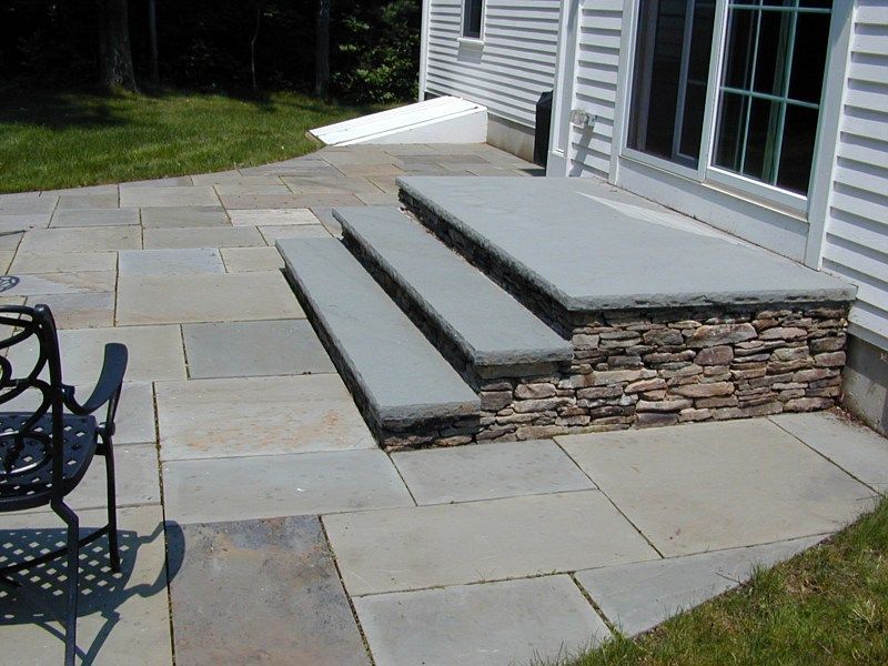 A stone patio with three wide, grey stone steps leading up to a sliding glass door on a white house exterior.