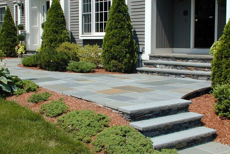 A stone walkway with steps leading to the front entrance of a house, surrounded by mulched garden beds and evergreen shrubs.
