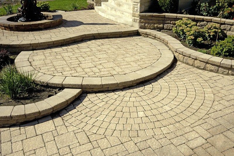 A tiered patio made of light-colored, interlocking concrete pavers featuring curved steps and a circular stone design.