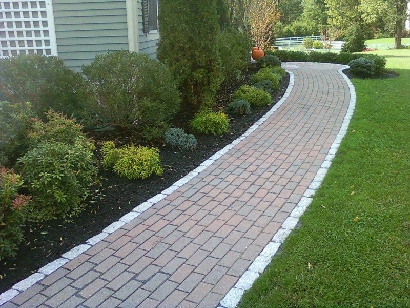 A curved brick walkway with white stone edging, bordered by landscaping shrubs and a green lawn next to a house.